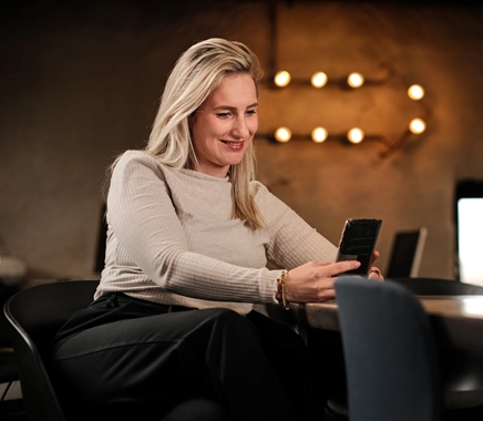 Jonge vrouw, met kort blond haar zit op een stoel en kijkt lachend naar haar telefoon