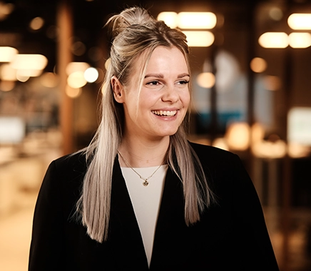 Jonge vrouw met lang blond haar kijkt naar rechts en lacht. Op de achtergrond een Persoonality vestiging