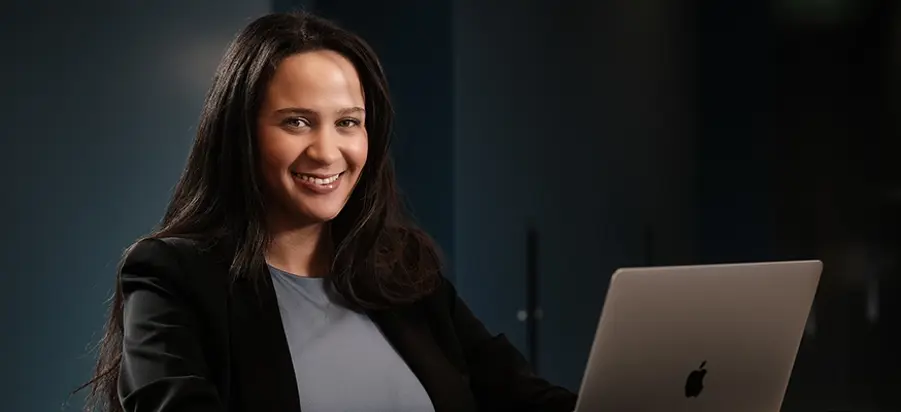 Jonge vrouw, met lang zwart haar zit achter de laptop, kijkt in de camera en lacht.